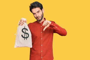 Young hispanic man holding dollars bag with angry face, negative sign showing dislike with thumbs...