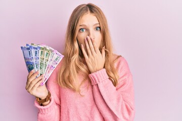 Beautiful young caucasian girl holding indian rupee banknotes covering mouth with hand, shocked and...