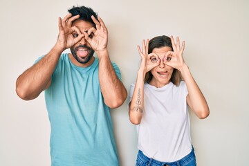 Beautiful young couple of boyfriend and girlfriend together doing ok gesture like binoculars sticking tongue out, eyes looking through fingers. crazy expression.