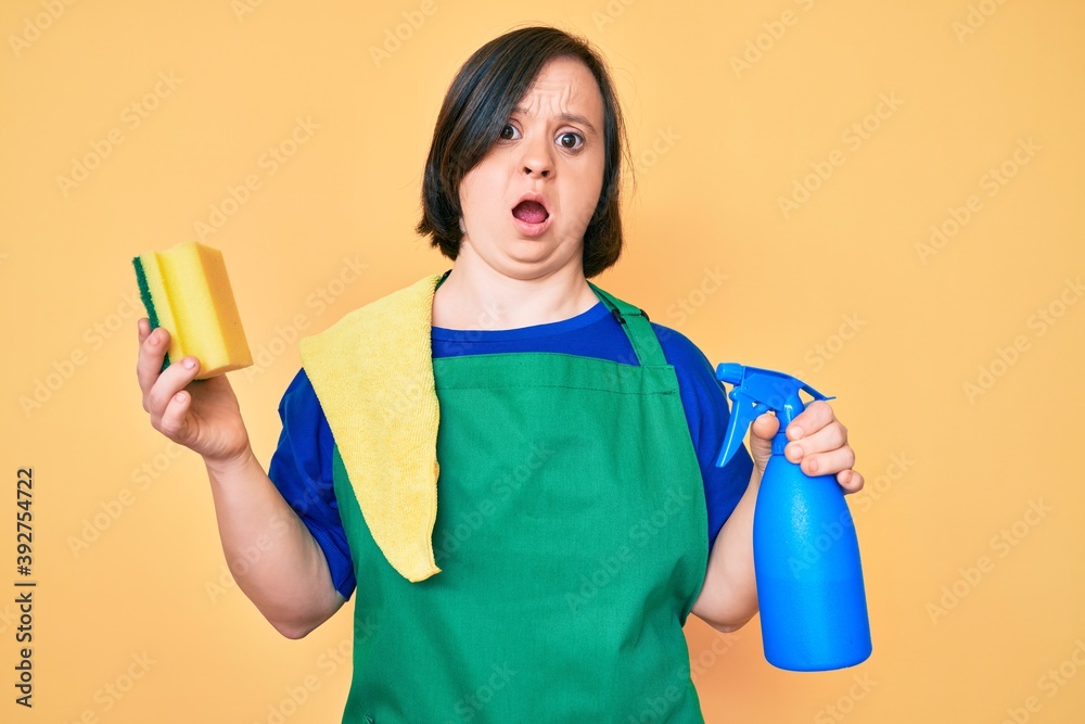 Sticker Brunette woman with down syndrome wearing apron holding scourer in shock face, looking skeptical and sarcastic, surprised with open mouth