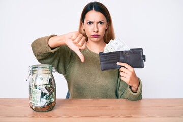 Young brunette woman with savings jar and wallet with dollars with angry face, negative sign...