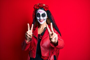Woman wearing day of the dead costume over red smiling looking to the camera showing fingers doing victory sign. number two.