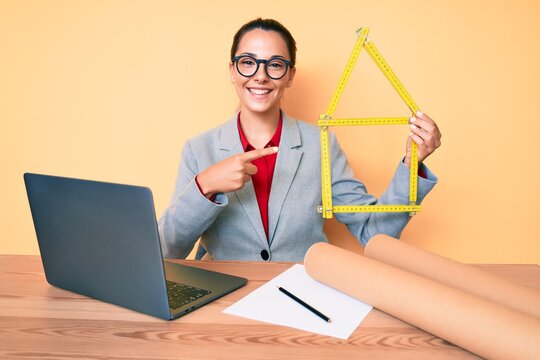 Young brunette architect woman at the office holding build project smiling happy pointing with hand and finger