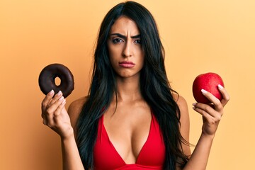 Beautiful hispanic woman holding red apple and donut skeptic and nervous, frowning upset because of problem. negative person.