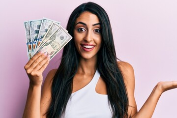 Beautiful hispanic woman holding dollars celebrating achievement with happy smile and winner expression with raised hand