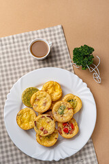 Egg bites with bacon, spinach and bell peppers on a round white plate on a napkin in a cell with a cup of coffee and parsley in a white basket on a beige background
