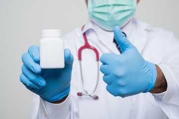 Close Up Asian bald Doctor in white coat, stethoscope, blue glove wearing face mask holding a bottle of pills isolated and get thumb up on white background.