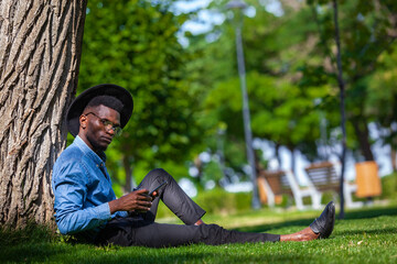 Obraz premium Portrait of a young black man in a hat and glasses sitting under tree in the park holding a cup of coffee in one hand and a mobile phone in the other