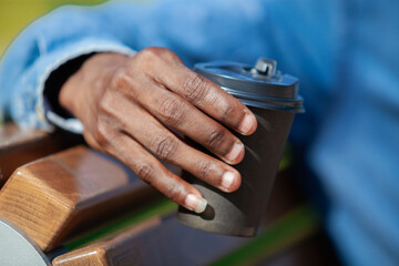 Close up of a coffee cup in a hand of black guy sitting on a bench in a park