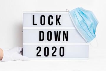 Hands in white gloves hold Light box with message lockdown 2020 and Surgical protective mask. Word...