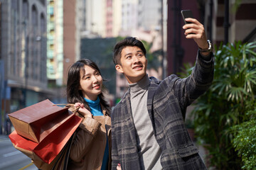 young asian couple taking a selfie using cellphone outdoors while shopping in the city