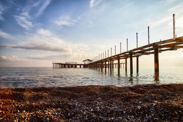 Obraz premium View from sand beach to water of sea, waves and pier in a nice day or evening with blues sky, bright sun and white clouds. The concept of a holiday on the sea or ocean in the South.