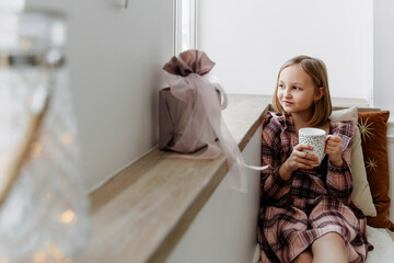 a young lady with a mug of tea sits on the windowsill. A ten-year-old girl looks out the window. Christmas concept