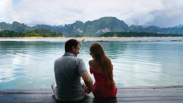 Romantic Pair of Man and Woman Sitting on Wooden Lake Pier with Green Mountains on Background. Funny Friends Fun and Laughing Together, Man Spanks Her Girlfriend, Flirting and Smiling