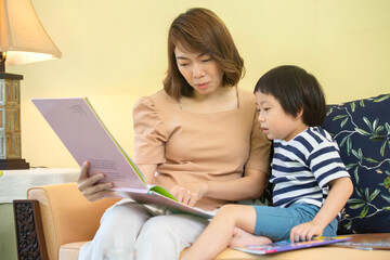 Asian mother is reading a storybook to her son on the sofa in the living room.