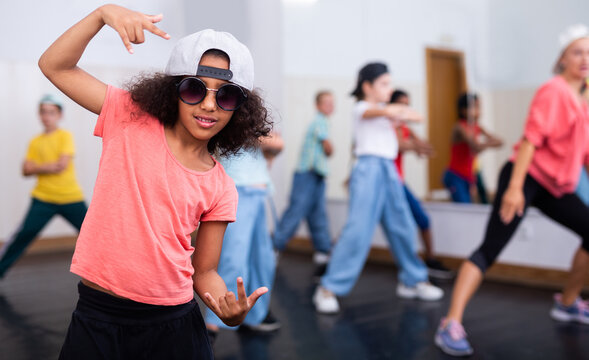 Confident preteen african american girl breakdancer in cap and sunglasses posing in dance studio with dancing children in background