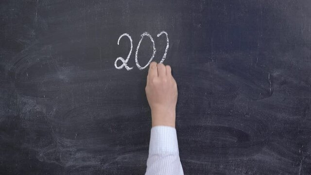 A hand writes 2021 in chalk on a black chalkboard and a question mark.