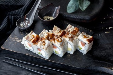 Uramaki roll inside out with smoked eel, salmon, cucumber and cream cheese. Unagi sauce with sesame seeds. Black background and black decor.