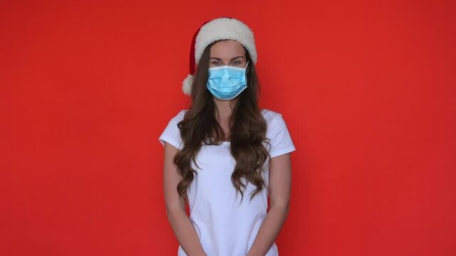 Woman in mask and new year hat waving hand. Portrait xmas woman waving hello in covid mask. New 2021 year in virus protection. Beauty model hello in orange studio
