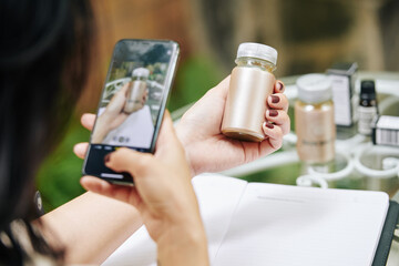 Close-up image of businesswoman taking photo of moisturising ampoule jar on smartphone for her online shop