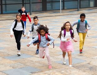 Multiethnic group of cheerful tweenagers running together in school yard after lessons in warm autumn day.