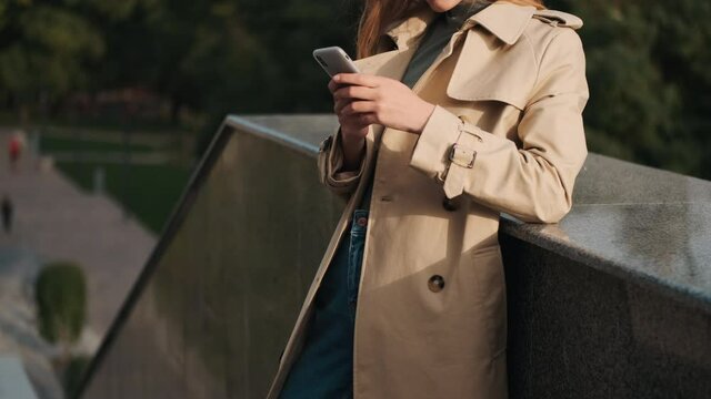 Conceptual shot of stylish woman dressed in trench coat using smartphone on street