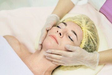 Fototapeta na wymiar Close up portrait of charming woman with closed eyes enjoying skincare procedure. Masseuse arms touching client chin and nose. Anti-aging effect