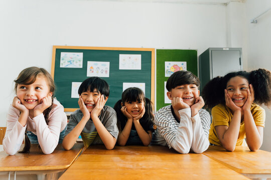 Cheerful multiethnic school friends facing camera with hands on chin, happiness, togetherness, friendship. Portrait of school children smiling in classroom