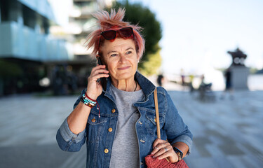 Senior woman with purple pink hair and smartphone walking outdoors in city or town.