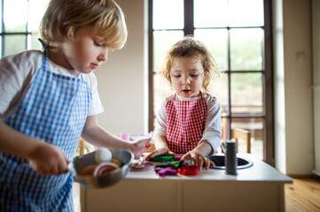 Small boy and girl with apron playing indoors with toy kitchen at home.