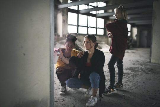 Group of teenagers girl gang indoors in abandoned building, using spray paint on wall.