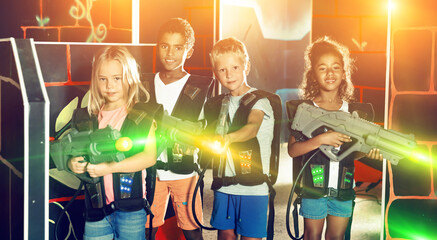 Cheerful tween girls and boys of different nationalities with laser pistols posing together at dark laser tag labyrinth
