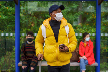Young man with smartphone on bus stop outdoors in town. Coronavirus concept.