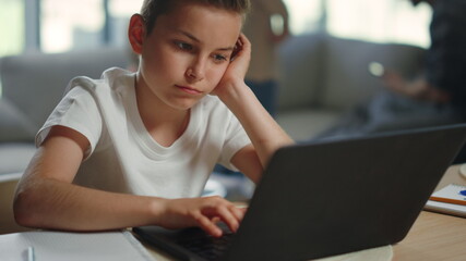 Sad kid studying remotely at home. Weary boy sitting at table with computer.
