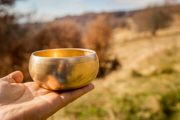Man hand holding a tibetan golden bowl against the nature background in the soft and beautiful light of morning. Yoga session, peaceful meditation music and positive energy through healing sounds.