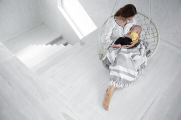 Happy mom feeds a baby in a white hammock with a blanket on the background of a white room. Happy motherhood concept.