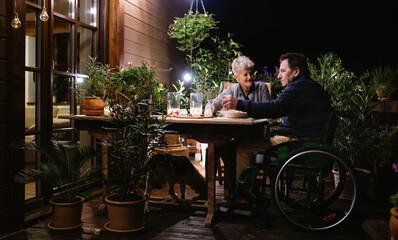 Senior couple in wheelchair having dinner in the evening on terrace, drinking wine.