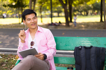 Asian man listening to music on a park bench in an outdoor.