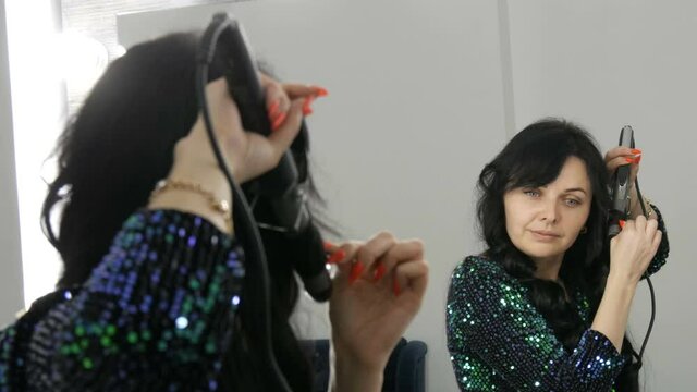 A beautiful spectacular young woman in front of a mirror winds her long black hair on a special iron to create curls.