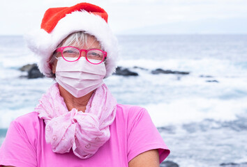 Serious senior woman wearing a protective mask due to the covid-19 coronavirus standing on the beach with the sea behind her. On his head a Santa hat while looking at the camera