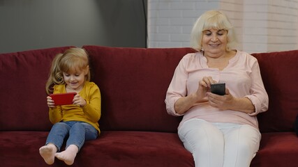 Senior grandmother with child girl granddaughter spending time home together, sitting on sofa in living room, using digital mobile phone. Watching videos, playing games, social network