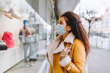 Beautiful afro haired woman wearing protective medical face mask stand on the street of city and look at shop window.