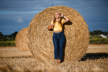 Naklejka na ściany i meble A young beautiful girl in a field stands near a sheaf. Vacation in the village. Young woman in yellow top and jeans