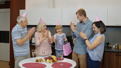 Loving mother, father and senior grandparents make surprise for cute child daughter and giving gift bag. Happy little kid girl celebrating birthday anniversary, having fun, dancing at home kitchen