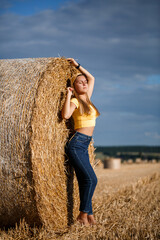 Naklejka na ściany i meble A young beautiful blonde stands on a mown wheat field near a huge sheaf of hay, enjoying nature. Nature in the village