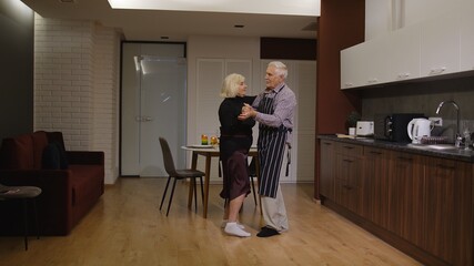 Happy senior couple in love have romantic evening, dancing together in the kitchen at home, celebrating anniversary. Elderly lovely husband and wife have supper with wine, in stylish cozy interior.