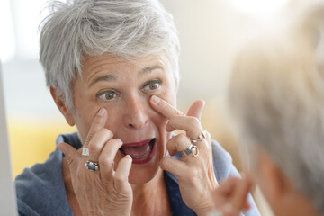 Mature white-haired woman checking eye wrinkles in front of mirror