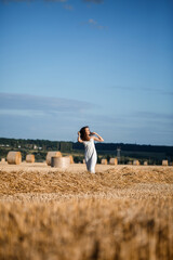 Naklejka na ściany i meble Young curly-haired woman in a wheat field, where there is a huge sheaf of hay, enjoying nature. People and travel. Nature. sun rays Agriculture
