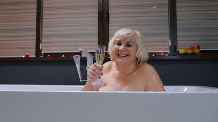 Sexy senior woman grandmother is taking foamy bath, drinking champagne in luxury bathroom with candles. Elderly lady grandma drinks white wine. Spa procedures self-care. Skin care. Life of retirees