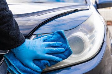 A hand in a blue rubber glove with a blue microfiber rag rubs and polishes the headlight of a car....